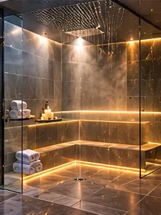 A glass-walled marble steam room with a rain shower, stone benches and rolled towels