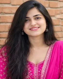 Meera in a magenta kurta, standing against a red brick wall