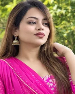 Riya in a bright pink saree, one hand in her hair, outdoors in soft light